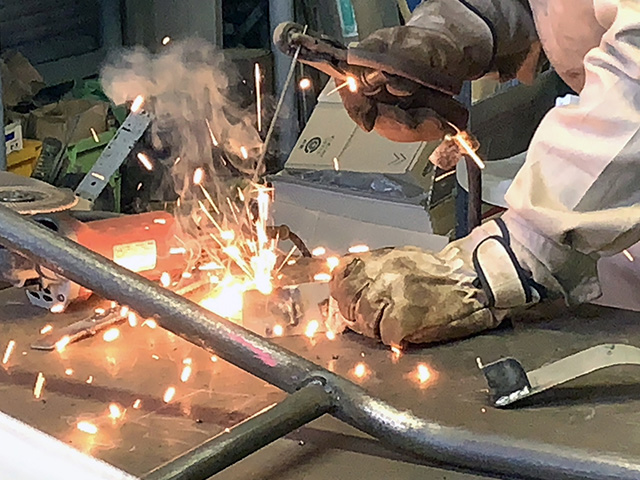 工場内で職人が溶接棒を使用し、金属の配管を溶接している様子。激しい火花が散り、周囲には資材や工具が置かれている。