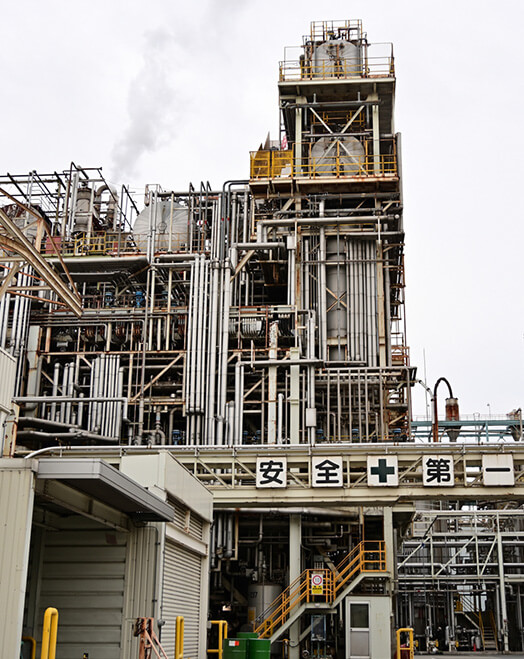 青空を背景にそびえ立つ、複雑に配管が組み合わされた巨大な化学プラントの設備外観。「安全第一」と書かれた看板が見える。