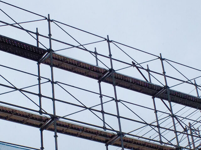 建設現場や修繕現場で高く組み上げられた、金属製の枠組み足場の見上げ写真。曇り空を背景に、安全通路となる足場板が設置されている。